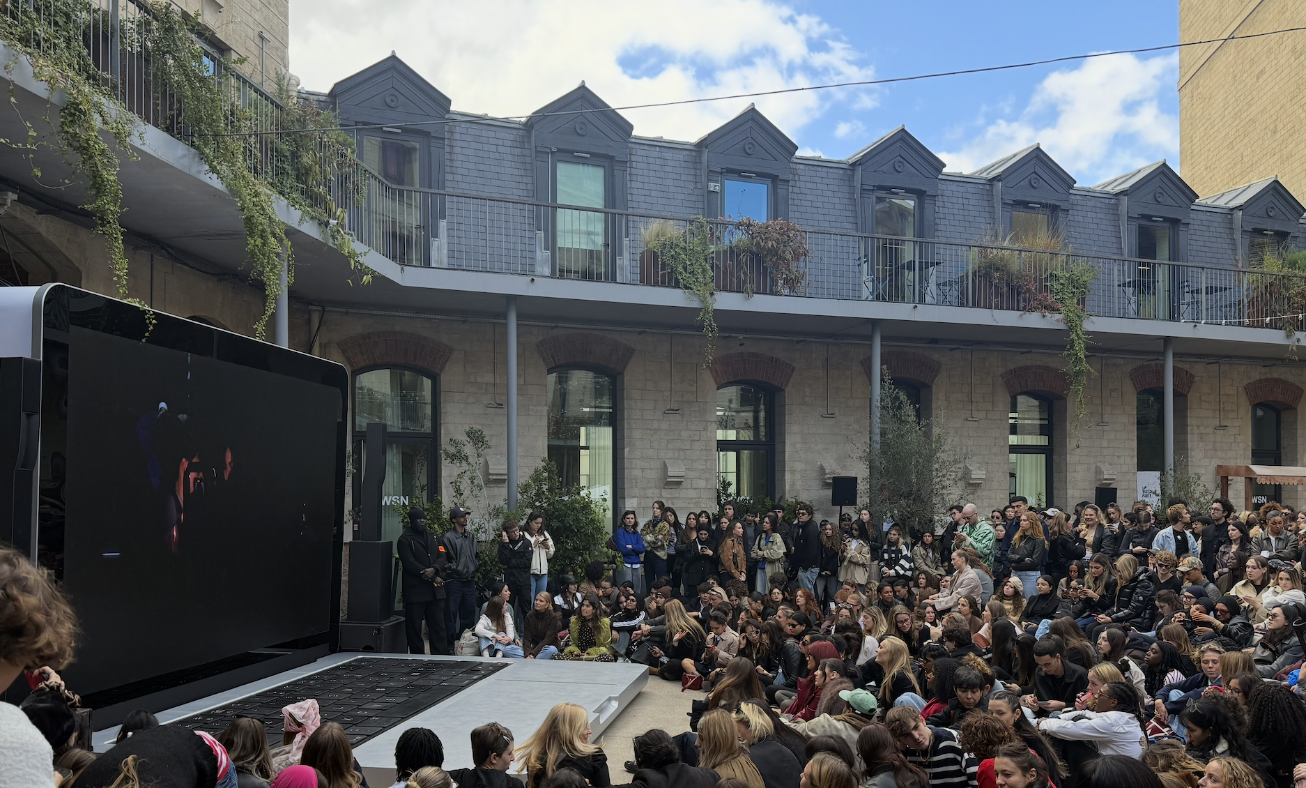 Parisian public square with a crowd of seated people watching a movie on a giant laptop