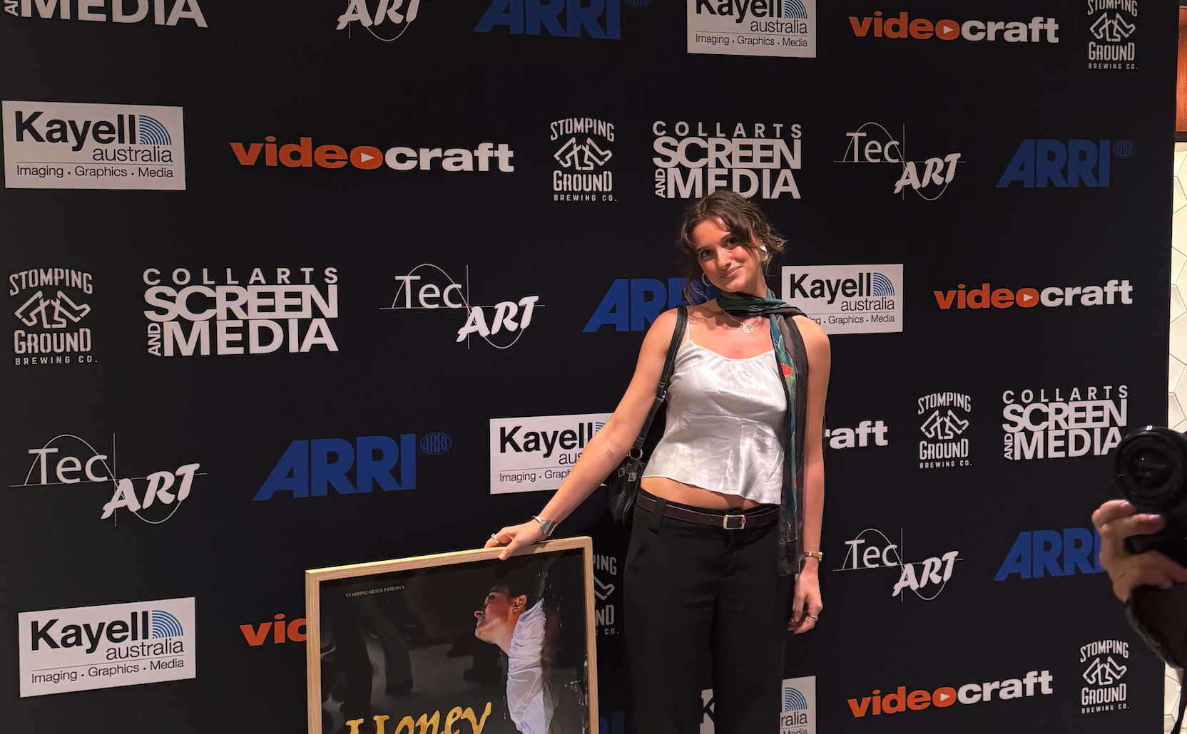 Collarts acting student posing with a framed filmed poster in front of a media wall with logos