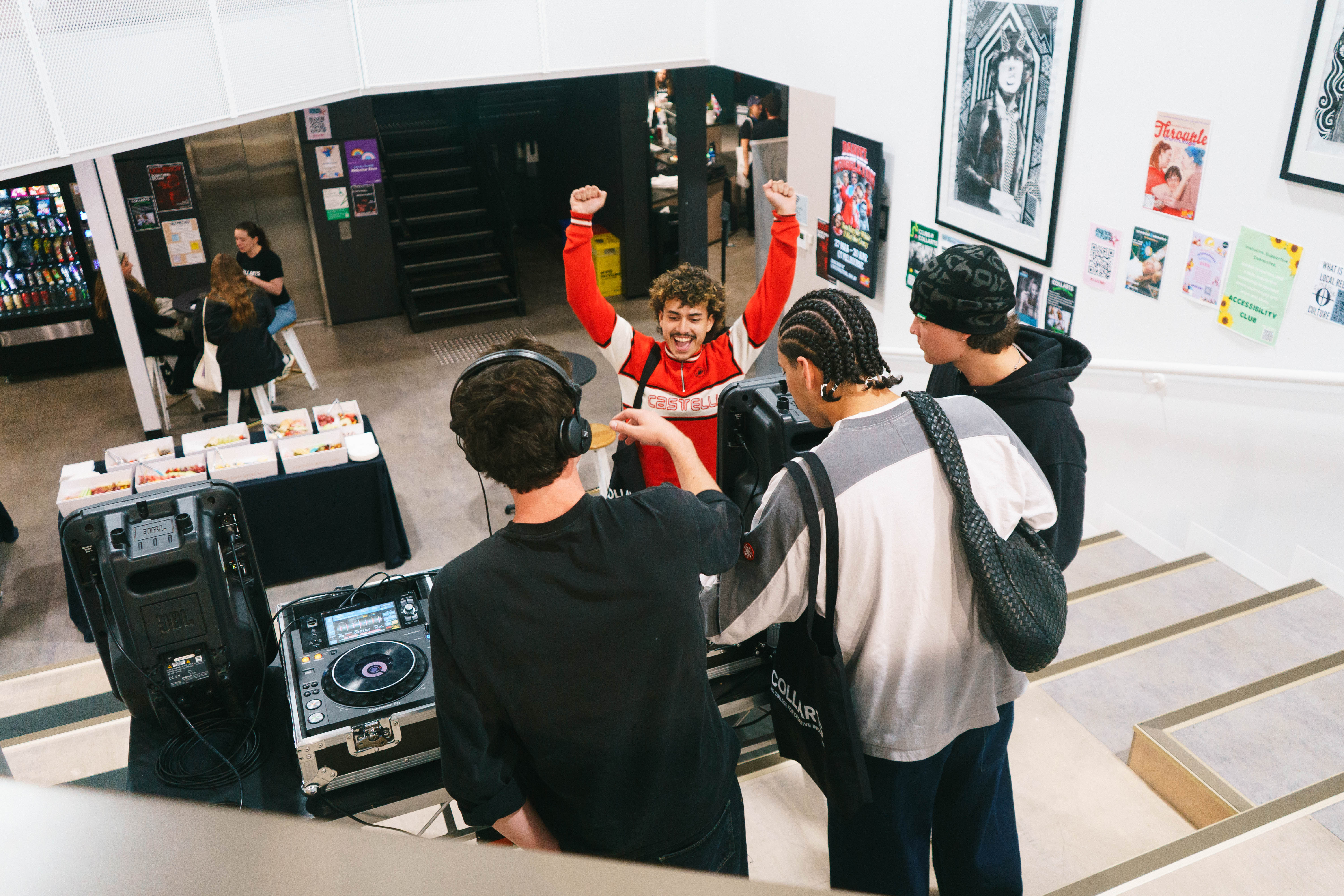 A group of music students gather around a DJ setup on a staircase, with one student wearing headphones while mixing music. Another music student in a red jacket throws their hands up excitedly, while two others watch and chat.  