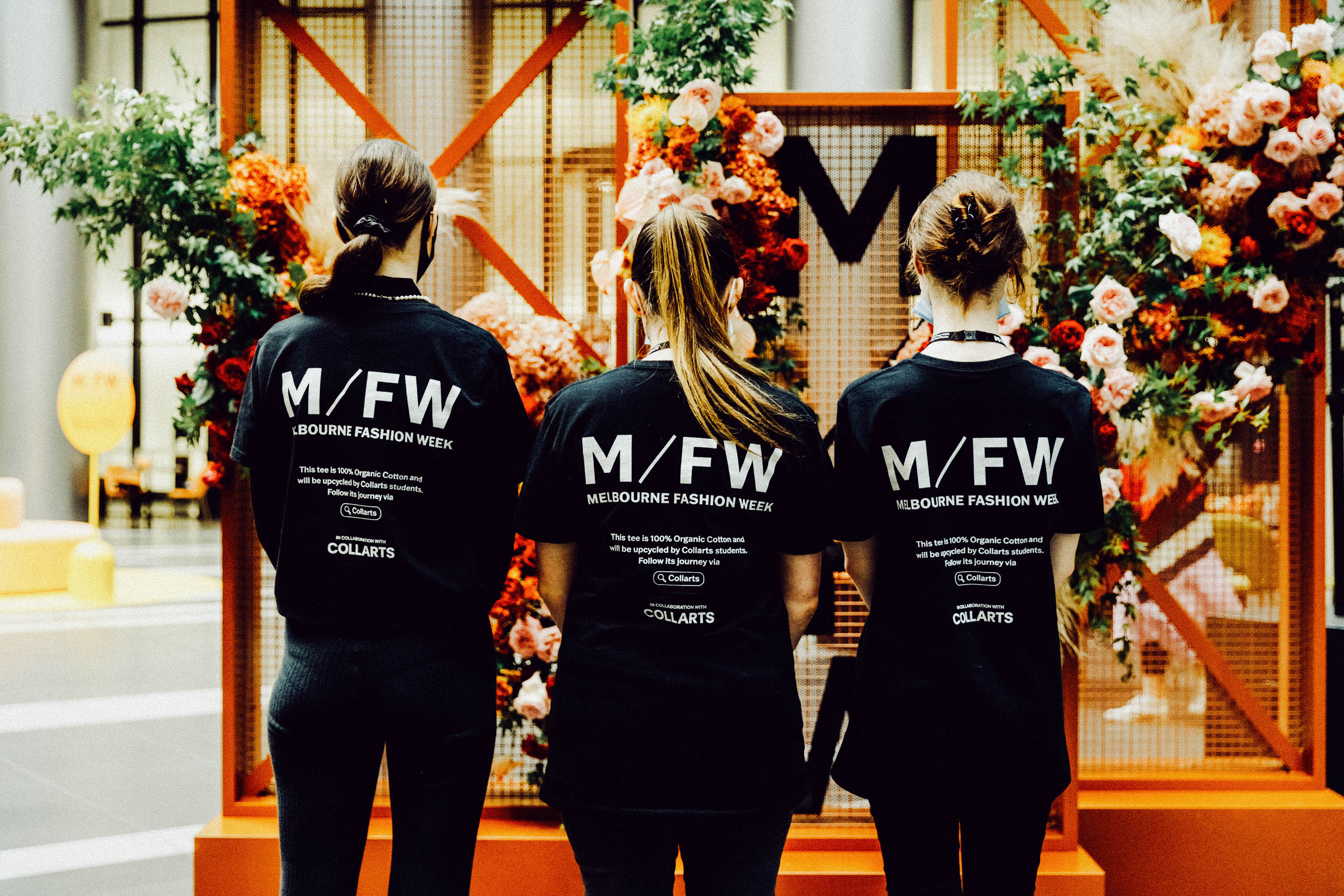 Three Collarts fashion students stand with their backs to the camera wearing black Melbourne Fashion Week x Collarts T-shirts. They are positioned in front of a vibrant floral display featuring red, pink, and orange flowers.
