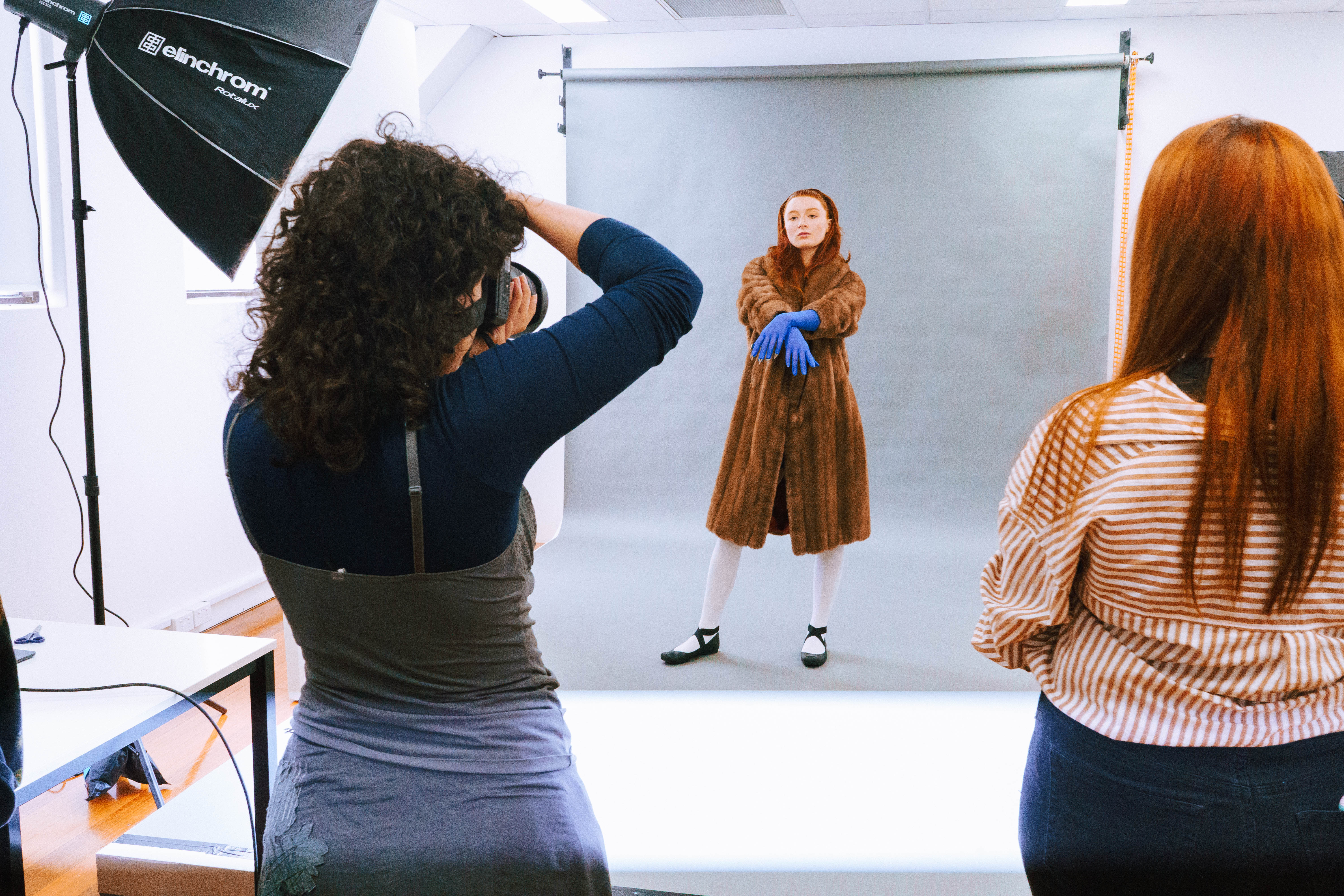 Fashion model posing with coat and gloves in studio while Collarts photography student takes photos of her